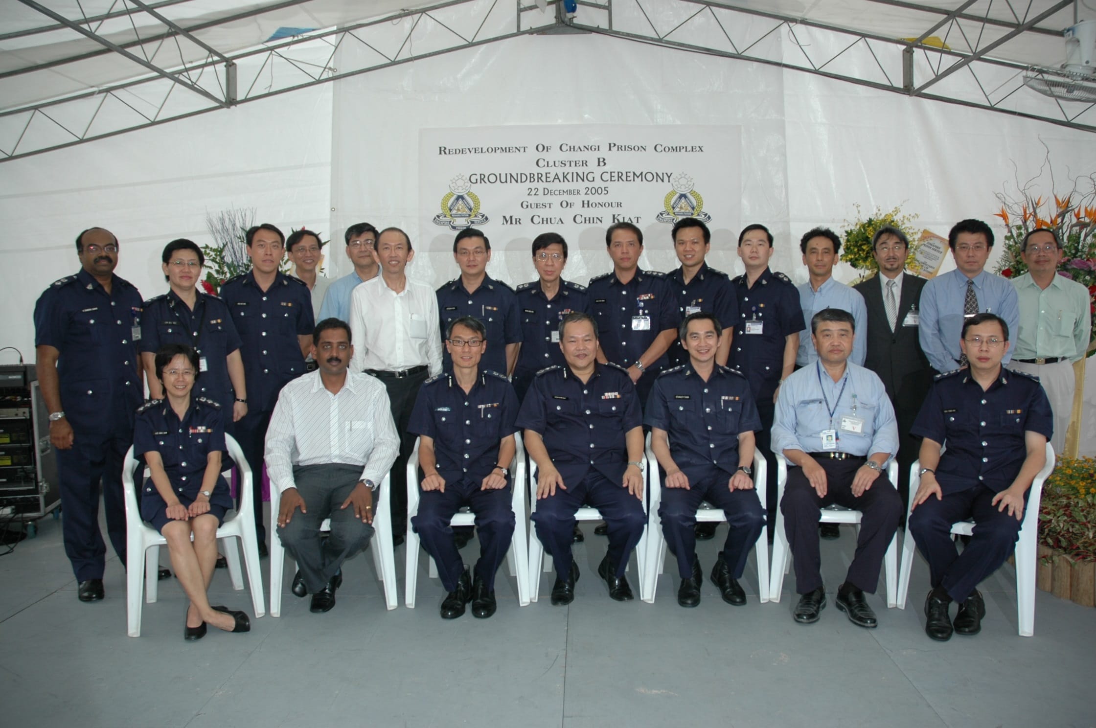 A group of people in uniforms and suits posing in front of a sign at a groundbreaking ceremony under a canopy.